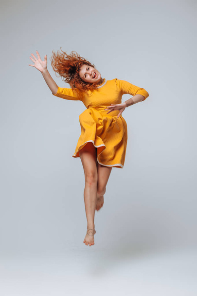 Red-haired girl in a yellow dress fun jumping on a gray backgrou - Foto, imagen