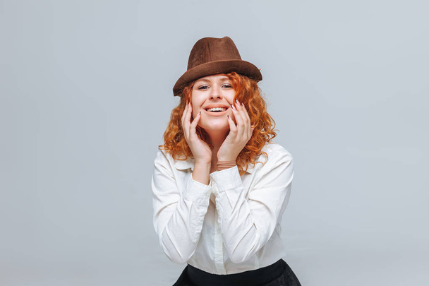 Červenovlasá radostná dívka v hnědém klobouku na šedém pozadí v  - Fotografie, Obrázek