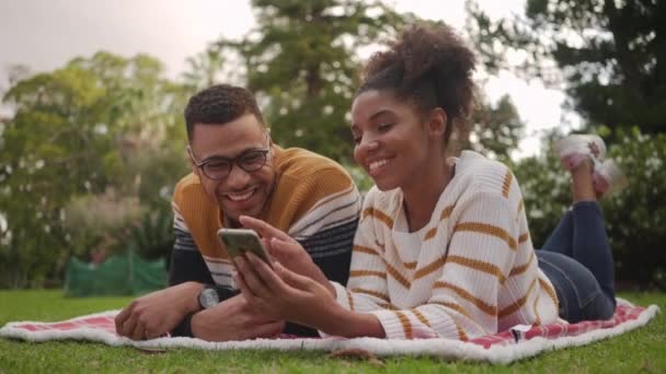 Relaxed smiling african young couple lying on blanket over the green grass taking selfie on smartphone in the park - Imágenes, Vídeo