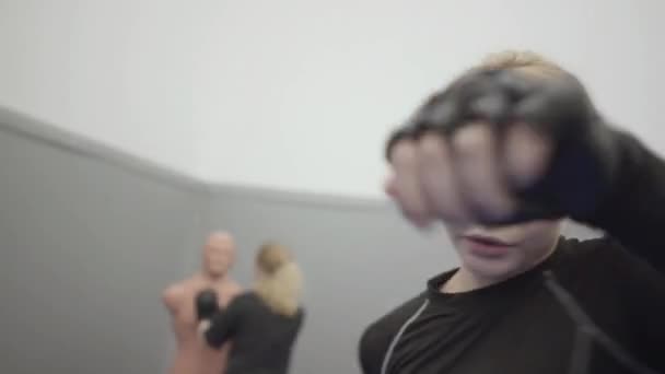 Jovem mulher loira bonita boxe na câmera em luvas de boxe preto. No fundo é mulher perfurando um manequim bob box
. - Filmagem, Vídeo