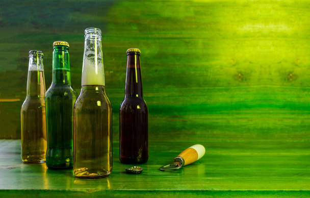 Group of beer bottles of different colors, one of them is uncovered and they are on a green textured wooden table - Foto, Imagem