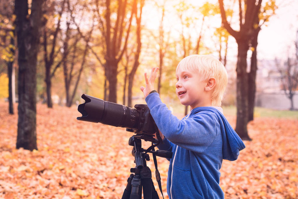 Kleine blonde jongen schiet met een grote SLR camera op een statief. Foto sessie in het herfst Park - Foto, afbeelding