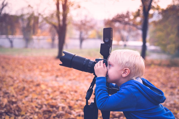 Küçük sarışın çocuk bir tripod üzerinde büyük bir SLR kamera ile vuruyor. Sonbahar parkında fotoğraf oturumu - Fotoğraf, Görsel