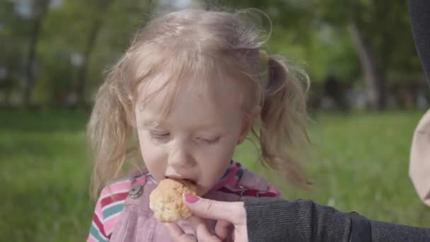 Hand of a young mother feeding her cute blond daughter in the park with a cookie close up. Active healthy lifestyle, connection with nature, leisure outdoors. Motherhood concept - Metraje, vídeo