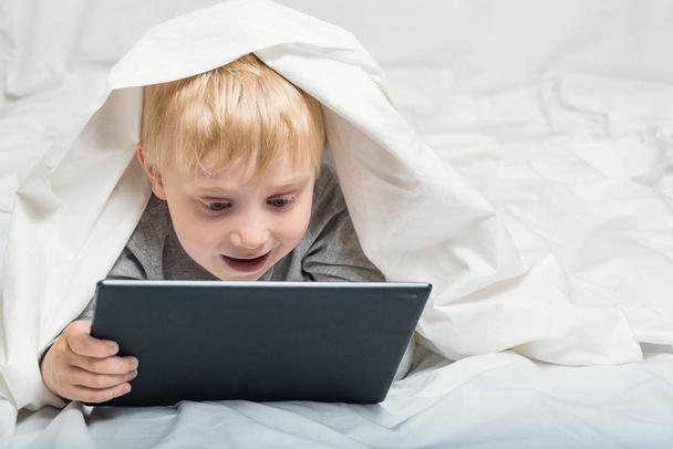 Kleine blonde jongen kijkt naar iets op een Tablet. Liggend in bed en ondergedoken onder de covers. Gadget Leisure - Foto, afbeelding