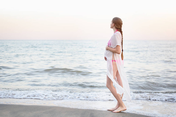 Молодая беременная женщина стоит на берегу моря и обнимает живот. Наслаждаясь моментом
. - Фото, изображение