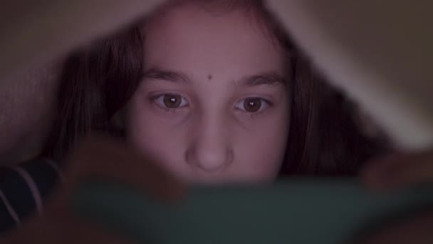 Aranyos tinilány, aki a takaró alatt heverészik a mobilján. Ideje lefeküdni. A kék fény negatív hatással van a gyermek szemére. - Felvétel, videó