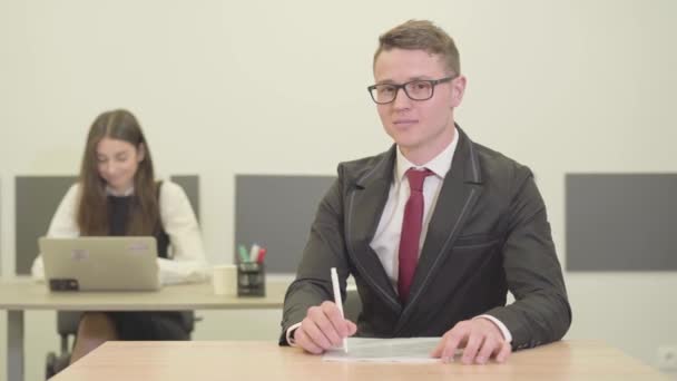 video of Confident young male manager in suit sitting at desk in foreground looking in camera. Attractive young woman in formal wear working with laptop on background - Video, Çekim