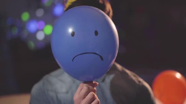 Portrait of an unhappy young man in birthday hat removing a blue balloon with a sad face painted on it from his head looking in the camera. The guy at the party, celebration concept - Кадри, відео
