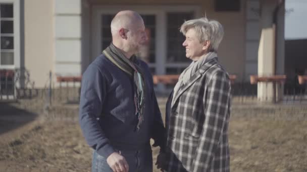 Happy senior couple, bald man and woman with grey short hair in coat and scarf stand near house and male showing a key in the hand. Bought new big house. - Footage, Video