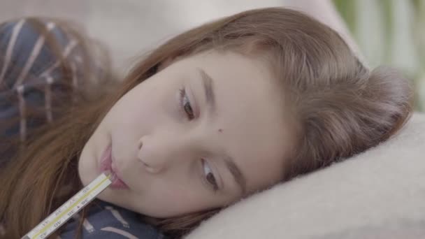 Sad tired sick girl lying in bed with a thermometer in mouth, video  - 映像、動画