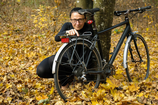 Eine 58-jährige Frau in schwarzer Kleidung und Brille sitzt auf einer gelben Blattoberfläche neben einem Fahrrad. Aktiver Lebensstil. - Foto, Bild