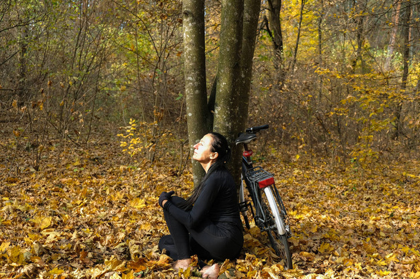 Femme aux pieds nus vêtue de vêtements noirs assise de profil sur une feuille jaune tombée près d'une bicyclette. Sur le visage souriant d'une femme les derniers rayons chauds du soleil. Concept d'auto-isolement. Changer la philosophie du concept de vie. - Photo, image