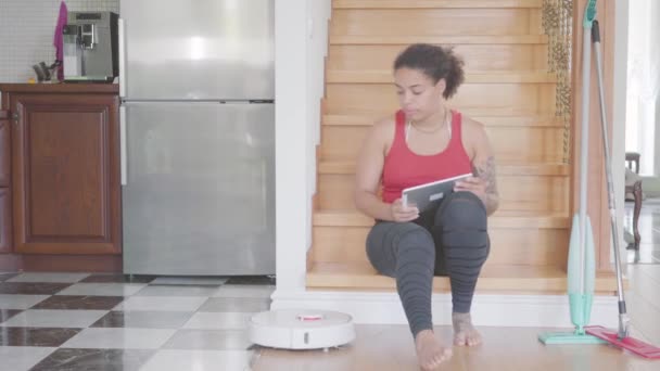 hübsche afrikanisch-amerikanische Frau sitzt auf der Treppe im ersten Stock eines großen Hauses und schreibt auf ihrem Tablet. moderner Roboter-Staubsauger  - Filmmaterial, Video