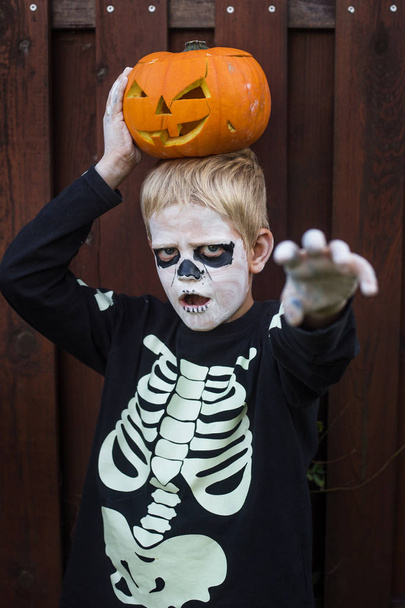 Happy young blond hair boy with skeleton costume holding jack o lantern. Halloween. Trick or treat. Outdoors portrait over wooden background - Photo, Image