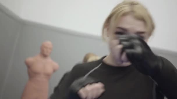 Fiatal, gyönyörű szőke nő bokszol a kamerában fekete boxkesztyűben. A háttérben egy nő üti a Bob Box próbababát.. - Felvétel, videó