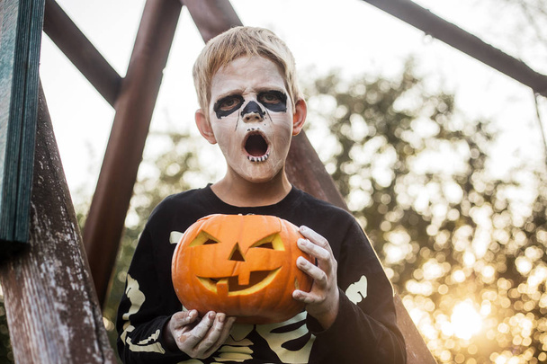 Happy young blond hair boy with skeleton costume holding jack o lantern. Halloween. Trick or treat. Outdoors portrait - Φωτογραφία, εικόνα