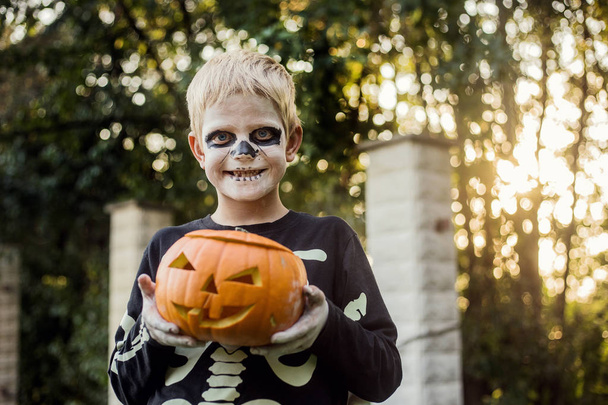 Happy young blond hair boy with skeleton costume holding jack o lantern. Halloween. Trick or treat. Outdoors portrait - Zdjęcie, obraz