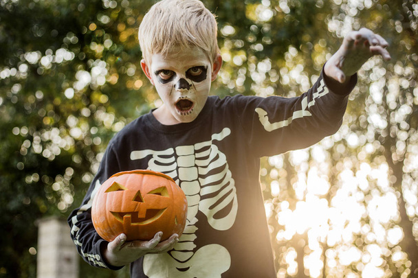 Happy young blond hair boy with skeleton costume holding jack o lantern. Halloween. Trick or treat. Outdoors portrait - Фото, зображення