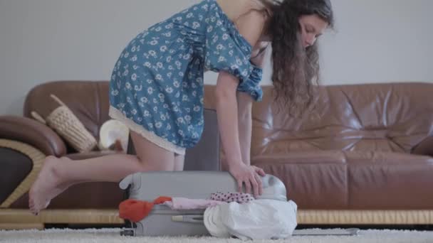 barefoot woman in dress trying to close suitcase sitting on it, Curly woman packing suitcase, video - Imágenes, Vídeo