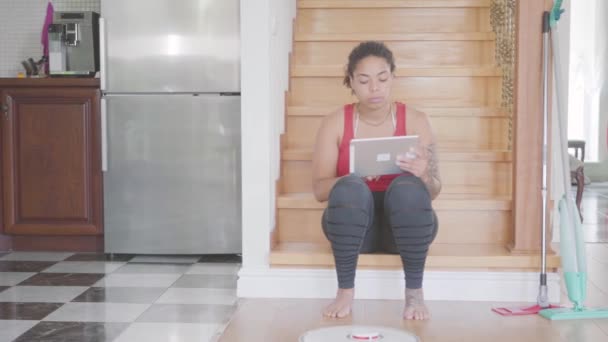 Video einer afrikanisch-amerikanischen Frau, die auf einer Treppe im ersten Stock eines großen Hauses sitzt und auf einem Tablet SMS schreibt. moderner Roboter-Staubsauger  - Filmmaterial, Video