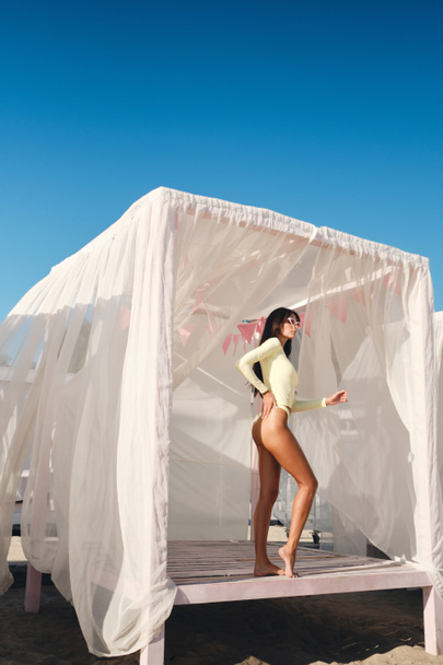 Attractive slim brunette girl in swimwear and sunglasses sunbathing on beach bed with curtains on seaside - Valokuva, kuva