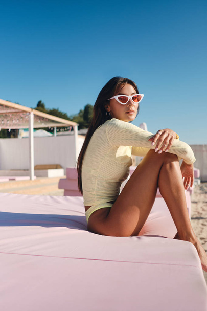 Belle fille brune élégante en maillot de bain et lunettes de soleil colorées regardant de rêve à la caméra sur lit de plage rose
  - Photo, image