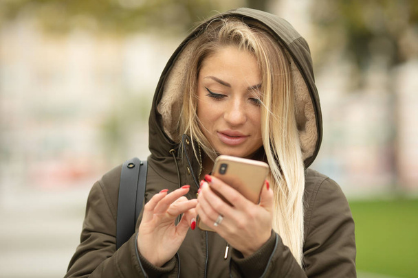 Portrait of young blonde woman in jacket using smartphone on urban background - 写真・画像