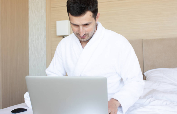 Young man in bathrobe using laptop in bedroom at morning - 写真・画像