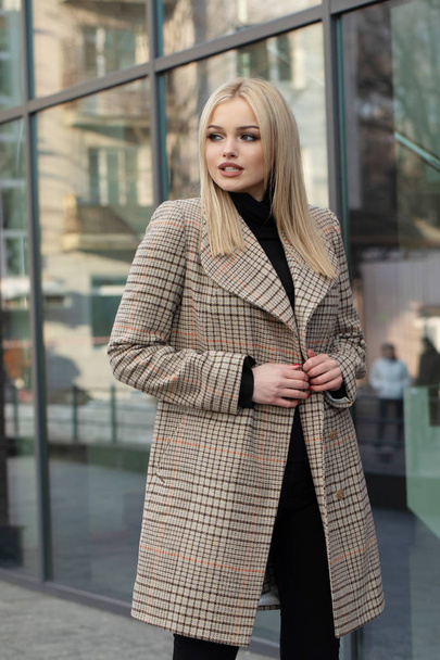 Blonde woman in warm beige woolen coat posing in city, modern building on background. - Fotó, kép