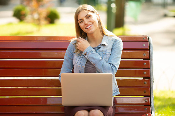 Young woman in denim jacket sitting on bench working with laptop in park - Zdjęcie, obraz