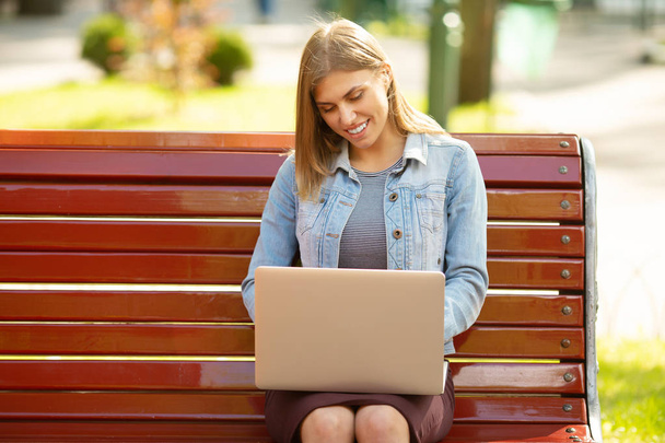 Young woman in denim jacket sitting on bench working with laptop in park - Фото, зображення