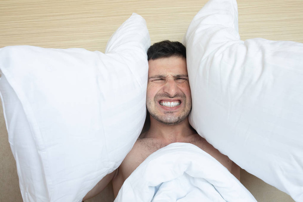 Homem irritado cobrindo orelhas com travesseiros sentados na cama de manhã
 - Foto, Imagem