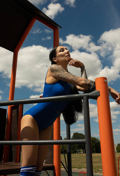 Fashion girl. Portrait of beautiful young tattooed hipster girl in blue swimsuit posing against urban background. - Foto, Imagem