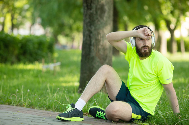 Young tired bearded man in headphones and sportswear resting after training in park - Φωτογραφία, εικόνα