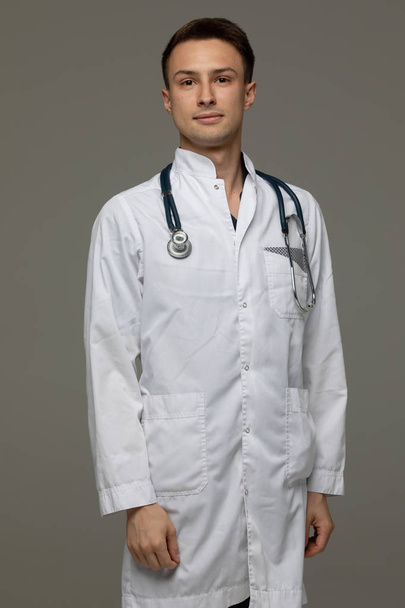 male doctor in white coat standing with stethoscope and looking at camera isolated on grey background. - Фото, изображение