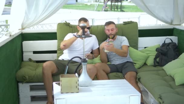 two friends smoke a hookah and watch the application on the phone while sitting in the gazebo outdoors on a sunny summer day. - Πλάνα, βίντεο