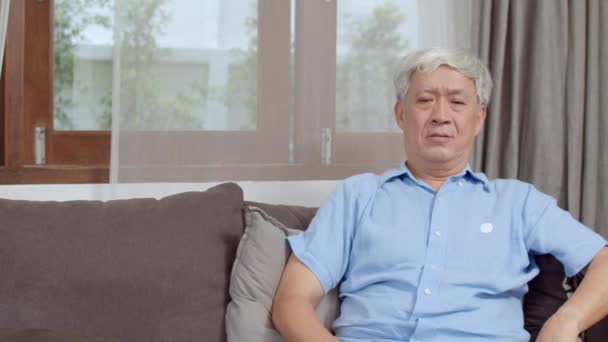 Asian senior man video call at home. Asian senior older Chinese male using mobile phone video call talking with family grandchild kids while lying on sofa in living room at home concept. - Séquence, vidéo