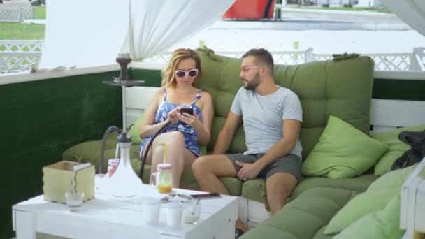 couple man and woman smoke a hookah in the gazebo on a clear sunny day and use a smartphone - Imágenes, Vídeo