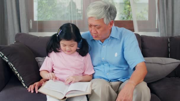 Ázsiai nagyapja pihenni otthon. Senior kínai, nagypapa boldog pihenni fiatal unokája lány élvezi olvasni könyveket és nem házi feladatot, valamint a nappali koncepció. Lassított lövés. - Felvétel, videó