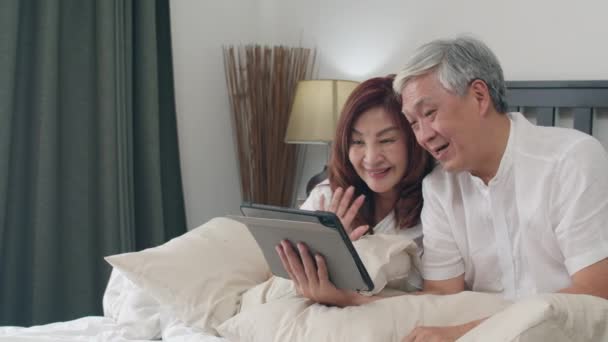Asian senior couple using tablet at home. Asian Senior Chinese grandparents, video call talking with family grandchild kids while lying on bed in bedroom at home in the morning concept. Slow motion. - Video, Çekim