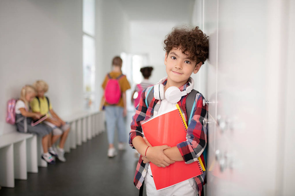 Cute curly schoolboy with earphones on neck standing near lockers - Photo, image