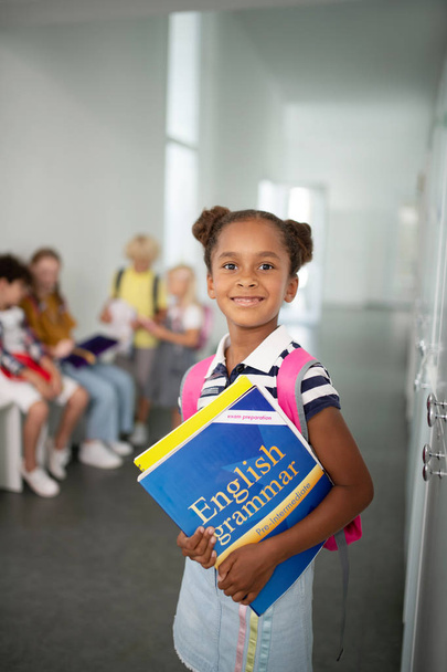 Nettes dunkelhäutiges Mädchen mit Büchern in der Hand, während es in der Schule steht - Foto, Bild