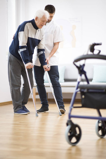 Elderly man walking on crutches and a helpful male nurse supporting him - 写真・画像