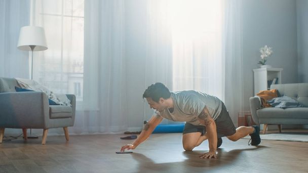 Athletic Fit Man in T-shirt and Shorts is Doing Mountain Climber Exercises While Using a Stopwatch on His Phone. He is Training at Home in His Bright Apartment with Minimalistic Interior. - Фото, зображення