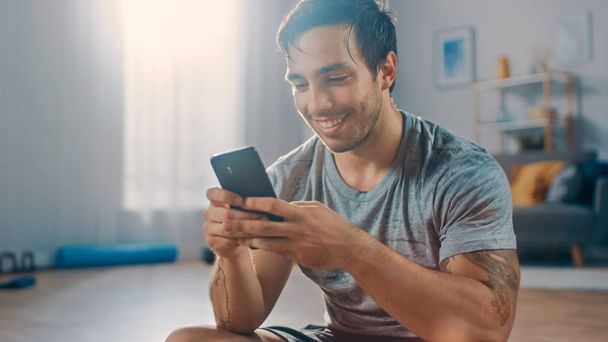 Happy Strong Athletic Fit Man in T-shirt and Shorts is Using a Mobile Phone After Morning Exercises at Home in His Spacious and Bright Living Room with Minimalistic Interior. - Foto, Imagen