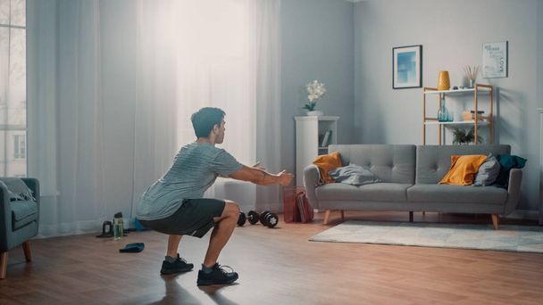 Strong Athletic Fit Man in T-shirt and Shorts is Doing Squat Exercises at Home in His Spacious and Bright Apartment with Minimalistic Interior. - Фото, изображение
