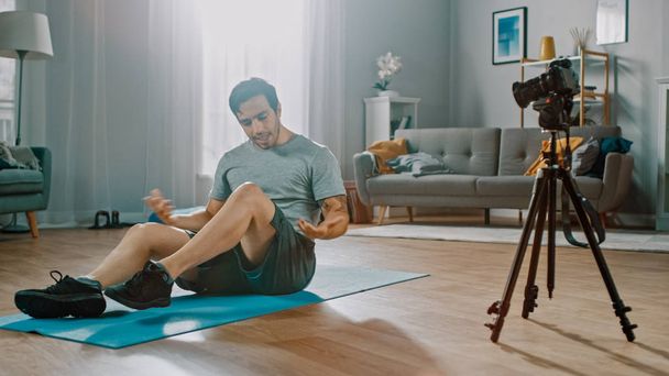 Strong Athletic Fit Man in T-shirt and Shorts is Recording his Workout on Camera for His Blog. Scene takes place in His Spacious and Bright Living Room with Minimalistic Interior. - Fotó, kép