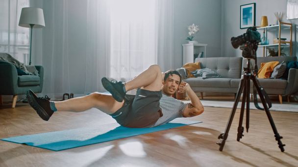Ein kräftiger, athletischer Mann in T-Shirt und Shorts hält sein Crisscross-Crunch-Workout für seinen Blog fest. Szene spielt in seinem geräumigen und hellen Wohnzimmer mit minimalistischem Interieur. - Foto, Bild