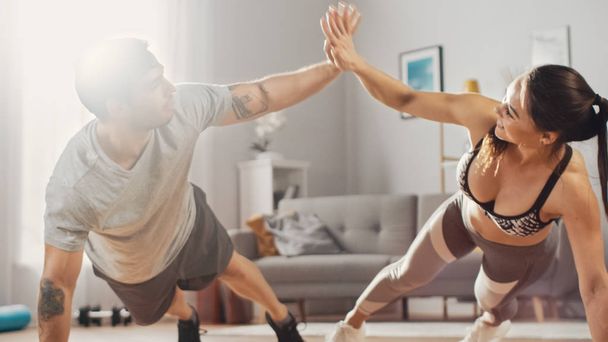 Strong and Beautiful Athletic Fitness Couple in Workout Clothes Doing Push Up Exercises and Giving Each Other a High Five in Their Bright and Spacious Living Room with Minimalistic Interior. - 写真・画像